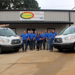 Lake Fork News A team of nine employees in matching blue shirts poses for a group photo outside the All Seasons Heating & Air Conditioning headquarters on Judson Road in Longview, Texas. The staff stands proudly before the gray building’s prominent company sign, with two branded white service vans displaying HVAC advertisements and roof-mounted ladders parked on either side, highlighting the local business’s presence in East Texas. Lake Fork, TX