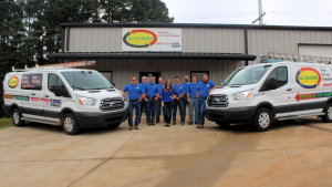 Lake Fork News A team of nine employees in matching blue shirts poses for a group photo outside the All Seasons Heating & Air Conditioning headquarters on Judson Road in Longview, Texas. The staff stands proudly before the gray building’s prominent company sign, with two branded white service vans displaying HVAC advertisements and roof-mounted ladders parked on either side, highlighting the local business’s presence in East Texas. Lake Fork, TX