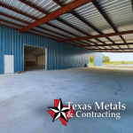 Lake Fork News Interior view of a spacious metal structure in East Texas, showcasing blue corrugated steel walls and a robust steel roof under natural daylight. Oversized doorways open to the rural landscape, capturing the distinctive “barndo”—barn-dominium—style popular in the region. The photograph emphasizes industrial craftsmanship and local architecture, with the Texas Metals & Contracting logo featured prominently at the lower right corner for clear attribution. Lake Fork, TX