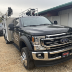Lake Fork News A black Ford Super Duty truck, outfitted for roadside assistance with utility storage compartments and emergency gear mounted on its roof, stands parked on a gravel lot outside a corrugated metal workshop in rural Northeast Texas. In the background, a second work truck is partially visible beside the main building under an overcast sky. Lake Fork, TX