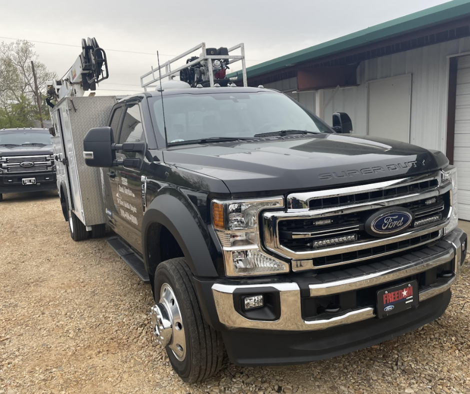 Lake Fork News A black Ford Super Duty truck, outfitted for roadside assistance with utility storage compartments and emergency gear mounted on its roof, stands parked on a gravel lot outside a corrugated metal workshop in rural Northeast Texas. In the background, a second work truck is partially visible beside the main building under an overcast sky. Lake Fork, TX